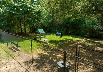The shady pet park features a bench, obstacles and grass.